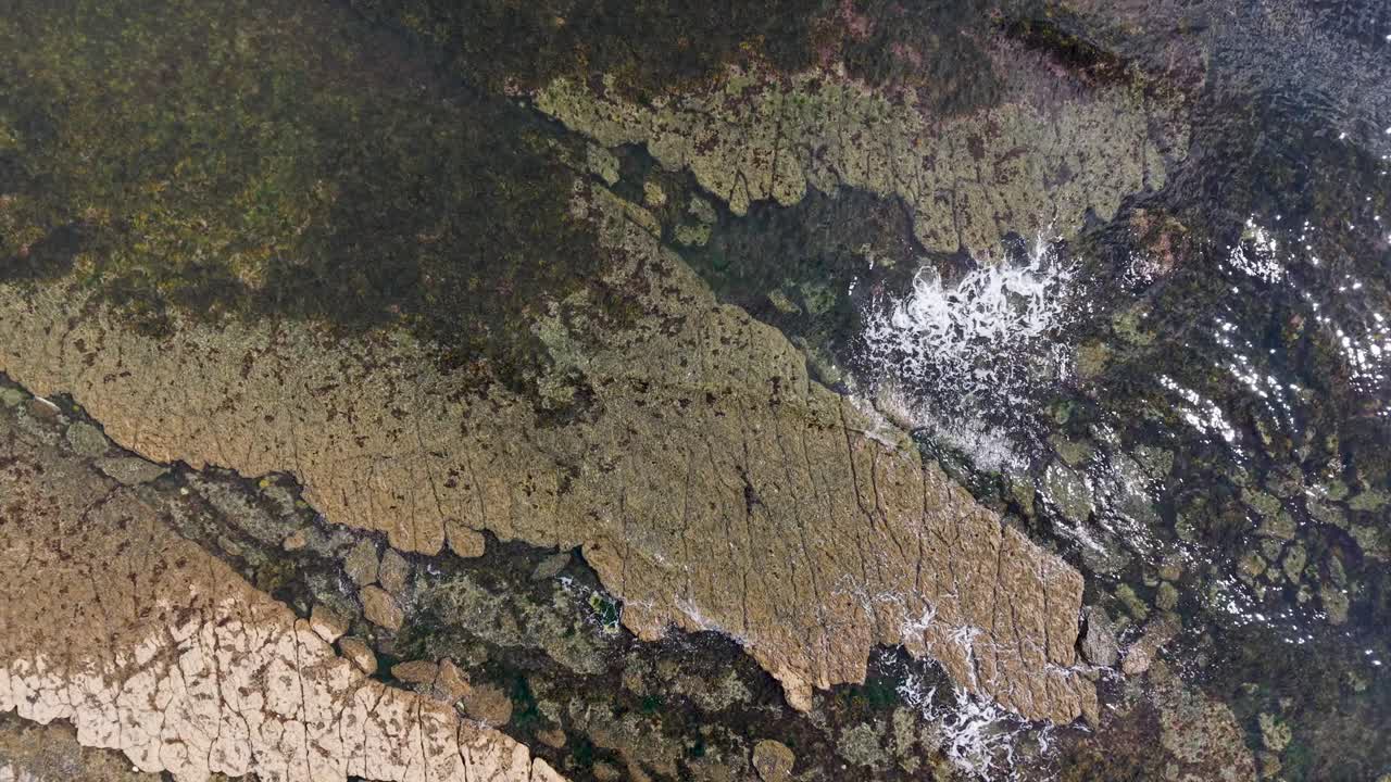 Aerial drone captures a top-down view of brown rocks in shallow sea. Waves crash gently with white foam forming as the drone lowers and slowly rotates, revealing textures of the rocky shoreline