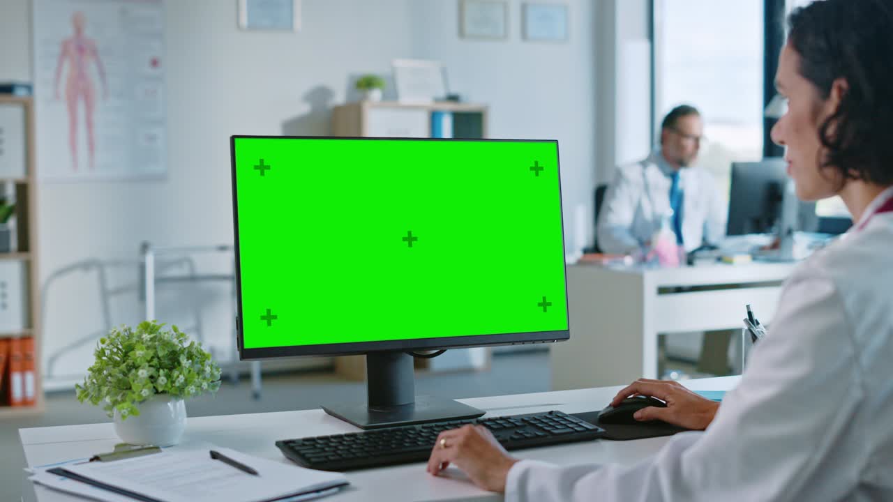 una dottoressa di medicina sta lavorando su un computer con schermo verde in una clinica sanitaria. un assistente in camicetta bianca sta leggendo la storia medica dietro una scrivania nell'ufficio dell'ospedale.