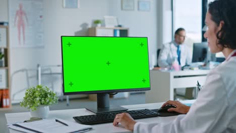 una mujer médica está trabajando en una computadora con una pantalla verde en una clínica de salud. una asistente con una bata blanca está leyendo historia médica detrás de un escritorio en la oficina del hospital.