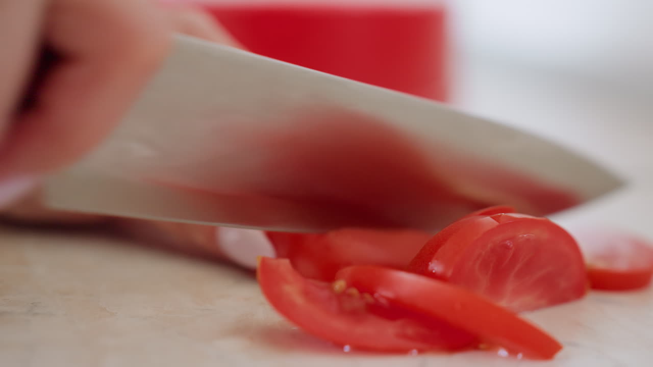Close up of cook slicing juicy tomato with sharp knife on kitchen counter, preparing fresh vegetable for healthy meal, highlighting texture of red slices and careful culinary process indoors