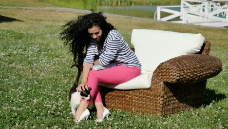 curly brunette relax in wicker armchair on green grass and strokes the cat