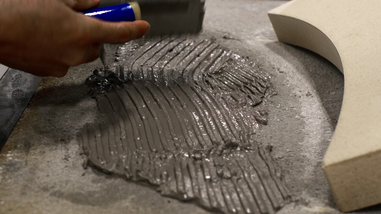 Close-up of mason spreading gray mortar with trowel onto oven base for curved refractory piece.