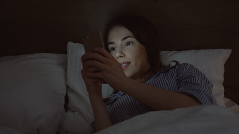 close-up view of the charming caucasian woman with dark hair lying in the bed, chatting on the smartphone late at night