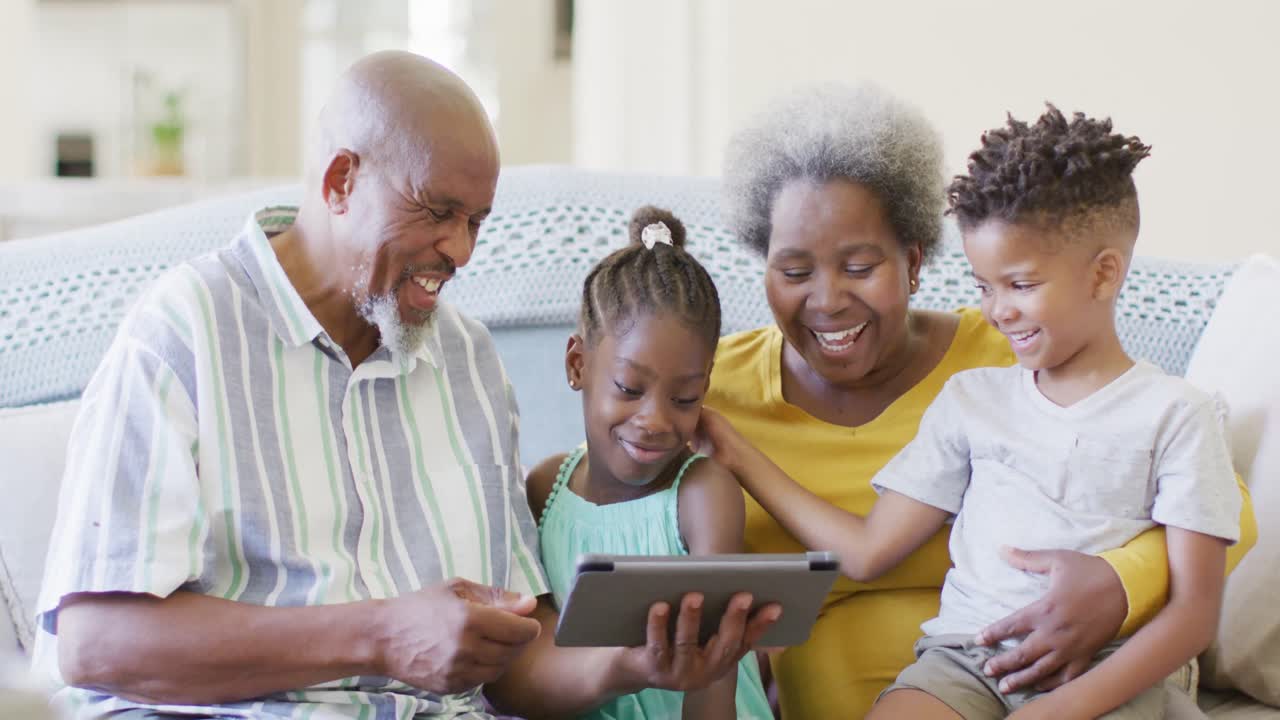 Happy african american grandparents, grandson and granddaughter on couch using tablet, slow motion