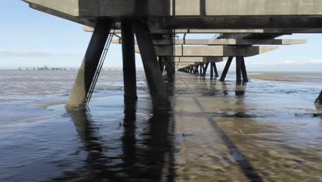 Fliegen-Unter-Betonpfeilern-Eines-Ölpiers-über-Der-Flachen-Küste-Der-Nordsee,-Deutschland