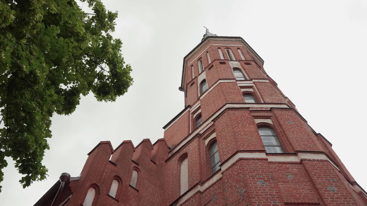 An upward view of a tall red brick church tower with gothic elements, framed by tree branches under a cloudy sky, showcases historical European religious architecture in Kaunas, Lithuania