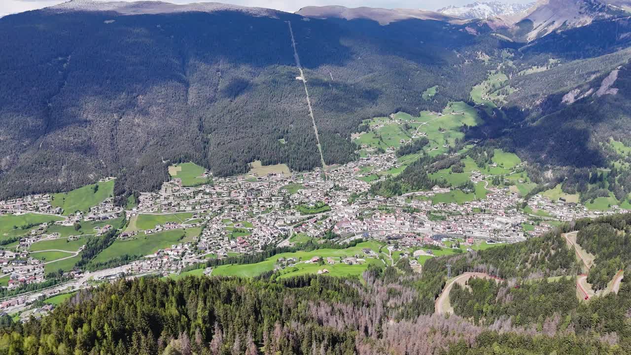 Ortisei in Val Gardena filmed by drone, dense forests, and snow-capped Dolomites mountains under a partly cloudy sky.