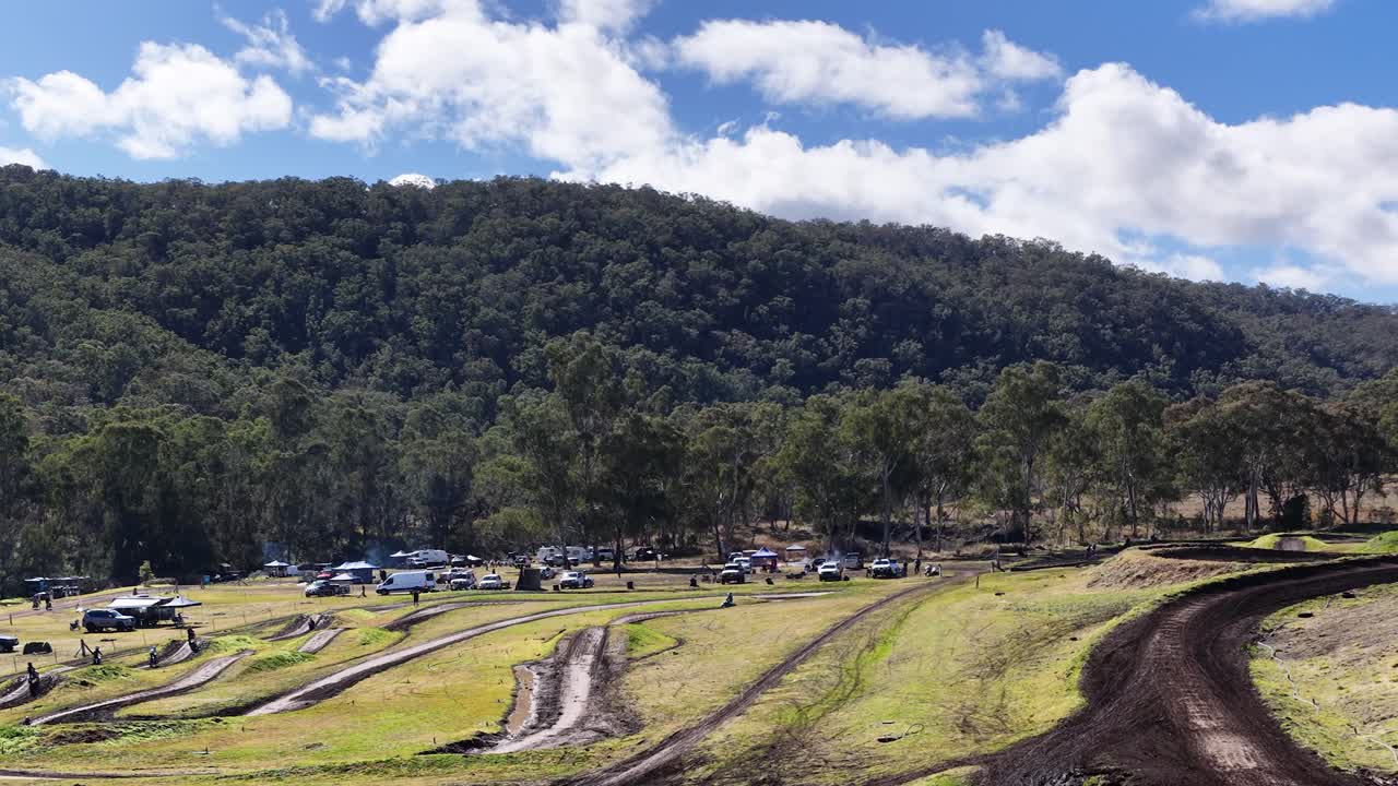 Motocross riders race on winding dirt track, surrounded by forested hills, under bright daylight