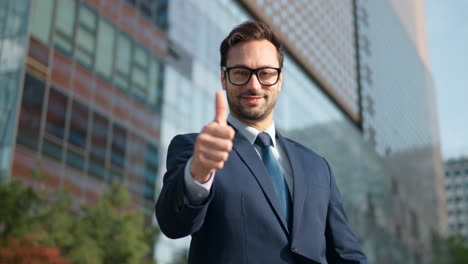 Successful-Business-man-in-a-suit-showing-big-thumb-up-outside-Glass-Building-Skyscrapers-at-sunset-with-big-smile