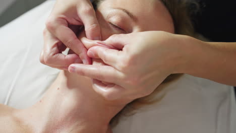 close up of relaxed female client reclined with eyes closed as beauty technician gently massages lips and surrounding cheek area with soft circular motions for soothing facial care