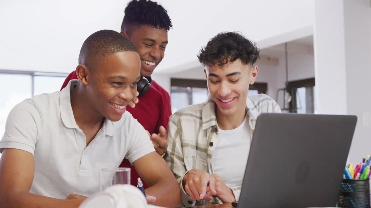 Three happy diverse male teenage friends using laptop and talking at home, slow motion