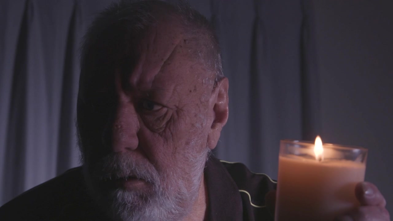 Um homem idoso olha pensativo enquanto segura uma vela acesa em um quarto escuro. A cena evoca contemplação e introspecção. Adequado para temas de solidão ou reflexão