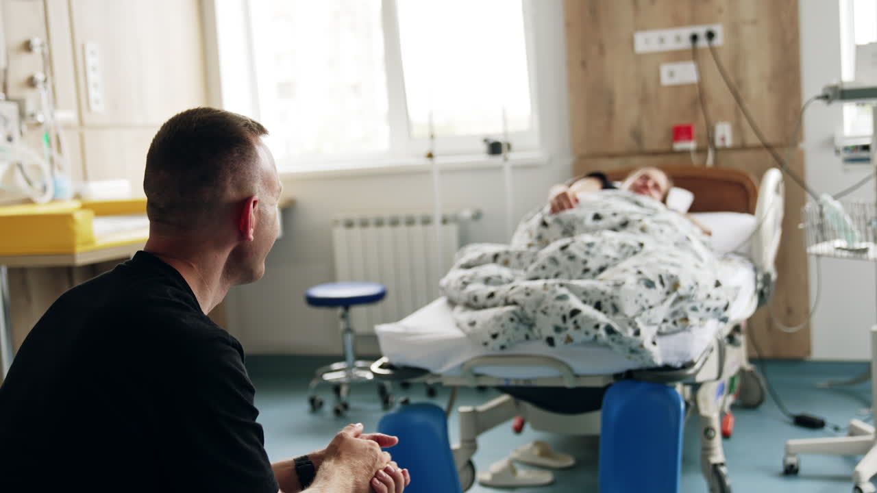 Rear view of a man sitting in the hospital ward. Husband looks at the bed where his pregnant wife lies. Blurred backdrop.