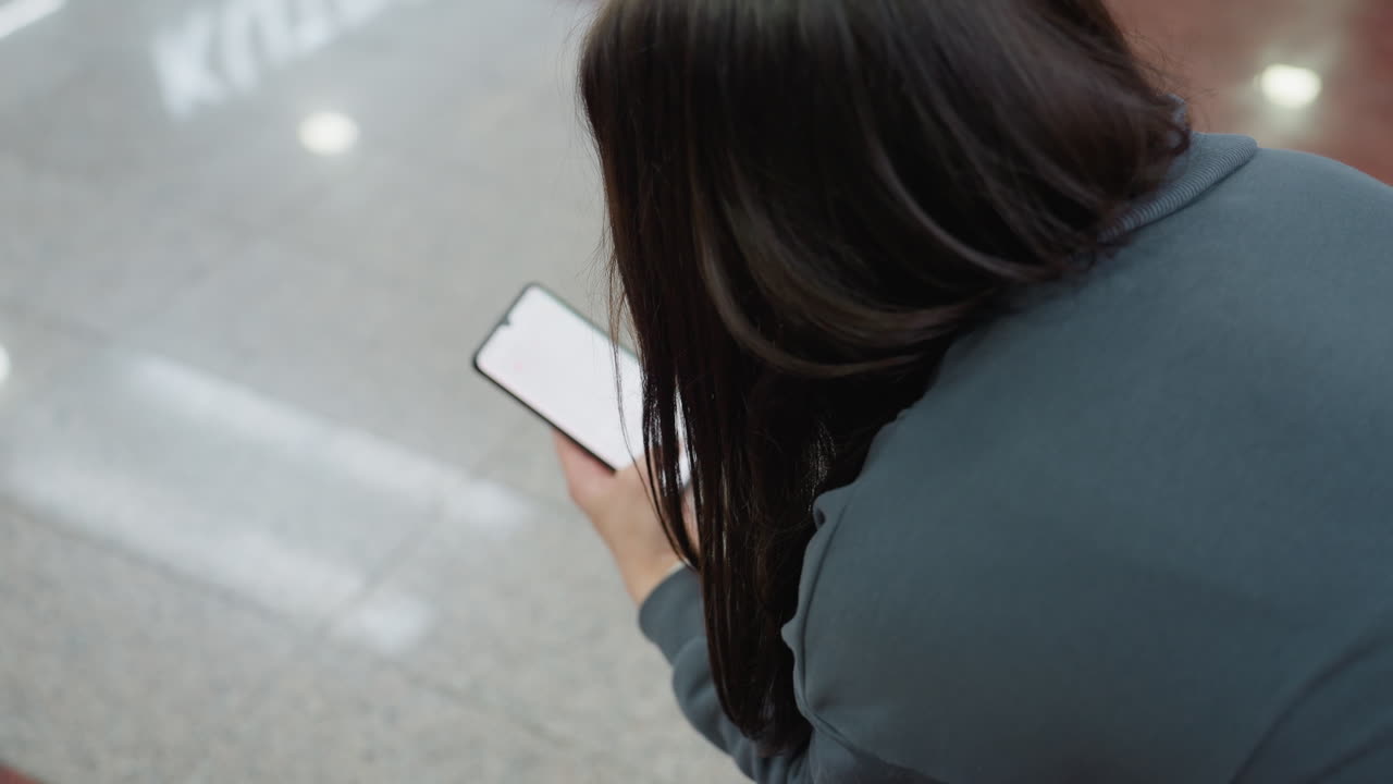 Rear view of lady messaging on phone while seated indoors, polished tile floor, blurred shoppers pass behind, casual dark outfit, long hair, urban mall mood of connection