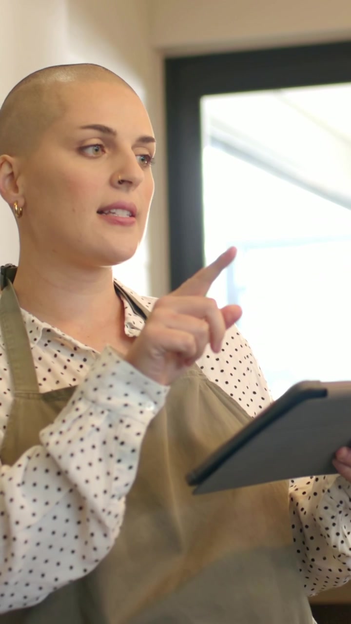 Vertical video: Reading adult woman explaining and pointing in kitchen with tablet and apron