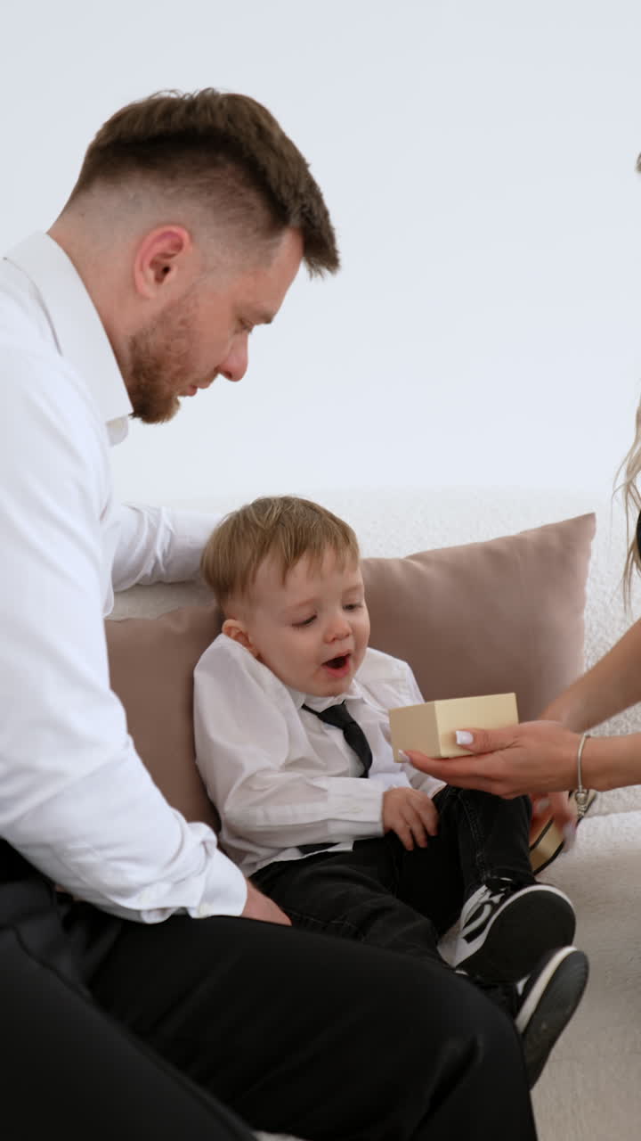 Family of three is sitting on the sofa. Mother is opening a little box in front of a kid. Giving a present to a child. Vertical video.