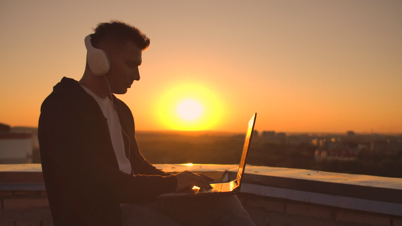 A man in a hoodie sits on the roof and listens to music with headphones typing on a laptop keyboard. Run at sunset. Freelancer works at sunset with a laptop