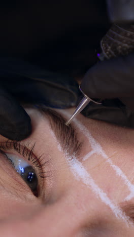 cosmetologist uses special tool to perform permanent eyebrow make up procedure. client's eyebrows are lined with white pen. close up. vertical video