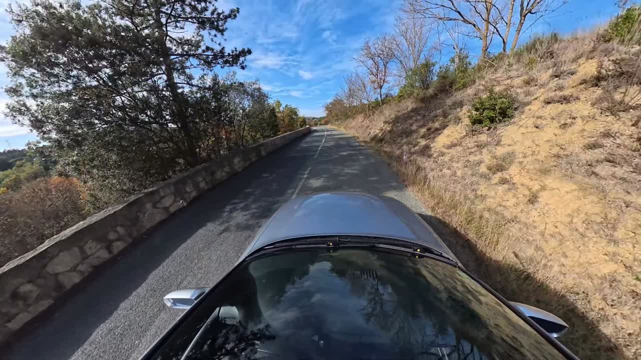 Aerial-style overhead shot of a silver car driving along a quiet countryside road on a sunny day. The footage shows the vehicle roof, windshield reflections, and scenic natural surroundings