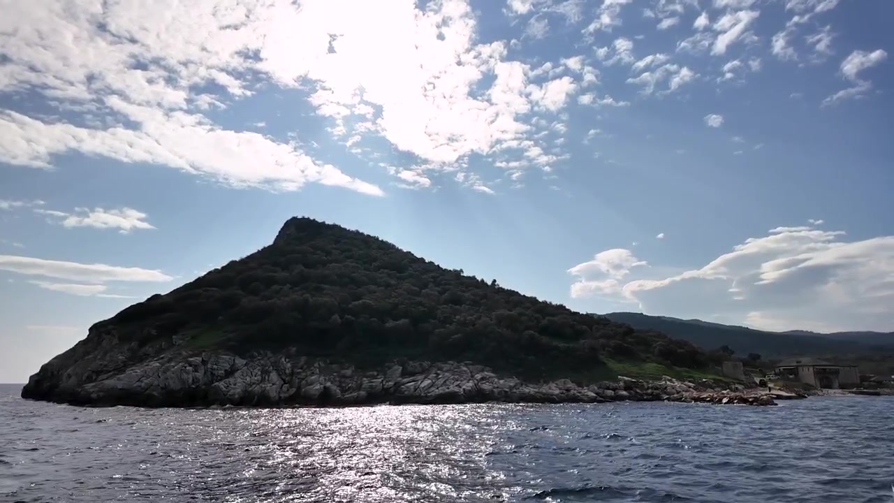 Ammira l'isola al largo della costa con colline verdi e coste rocciose, mentre il sole splende luminoso in alto. Le onde lambiscono dolcemente la riva, mentre il cielo mostra nuvole sparse.