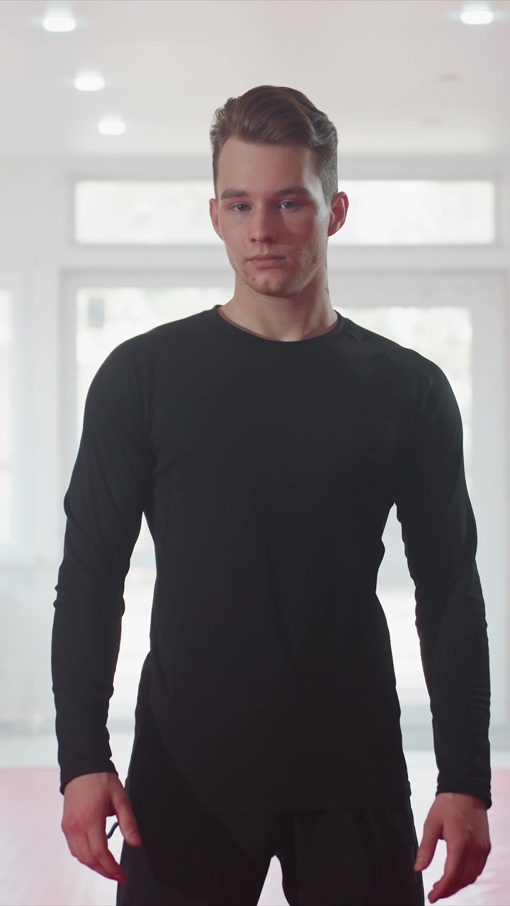 Young man with short brown hair and neck tattoo stands indoors. Man wears black long sleeve shirt in bright room with large windows. Camera remains stationary throughout focusing