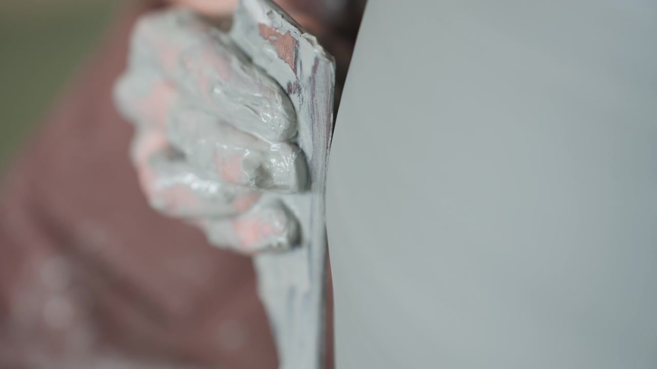 Gros plan sur des mains raclant la paroi d'un récipient dans un atelier de poterie, un potier affine l'épaisseur avec un petit outil, des éclats d'argile et une surface humide sont visibles, mouvement patient et précis pour un bord raffiné