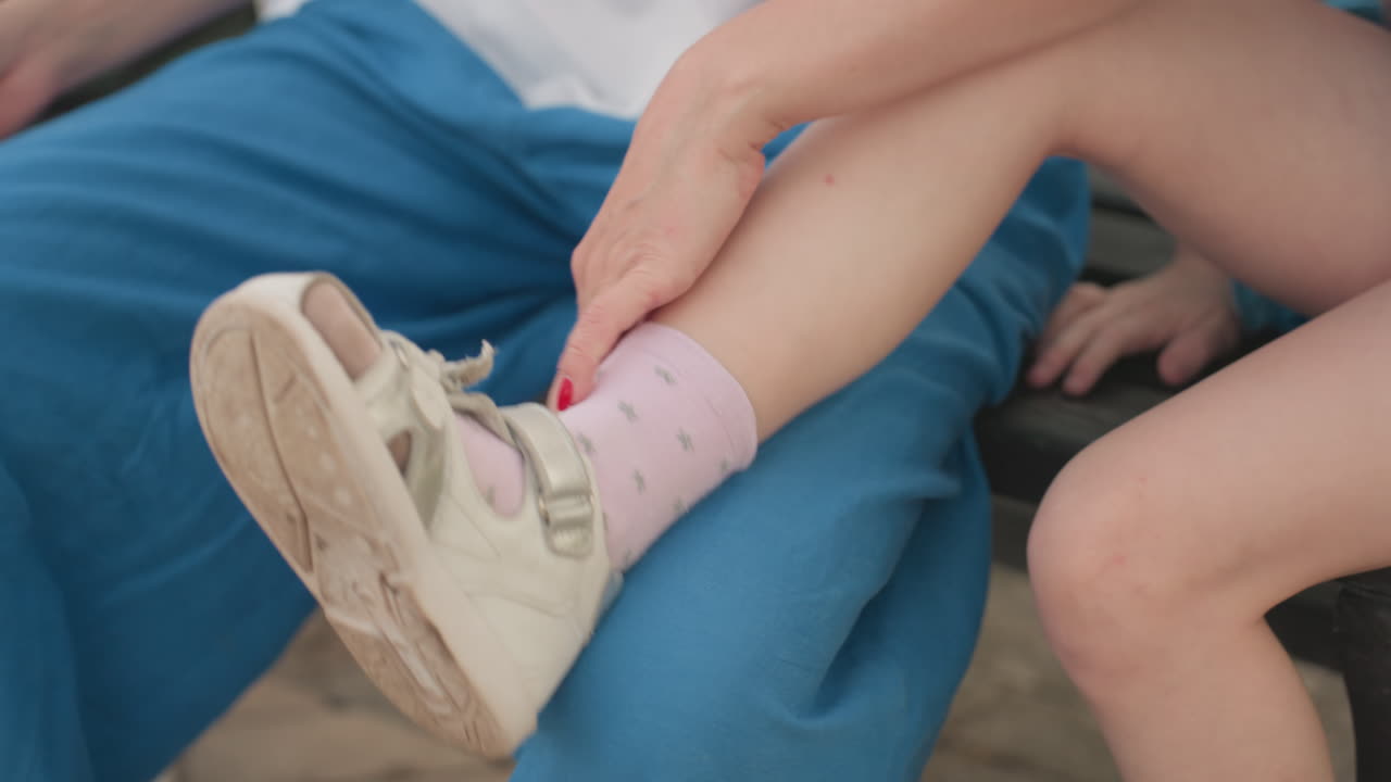 mother helping daughter put on small shoe while seated on bench in park, close up of hands adjusting strap, warm summer light, casual clothing, tender caregiving moment between parent and child