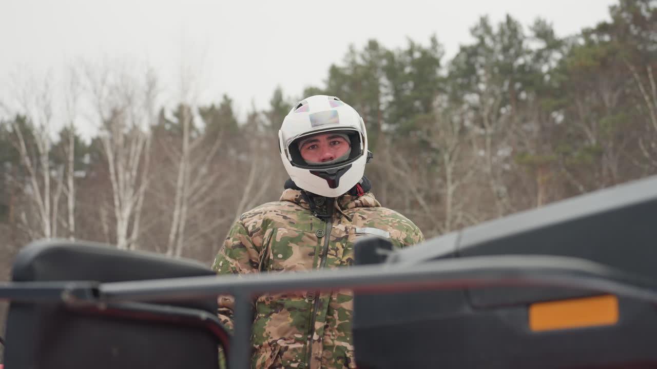 Pilota di ATV con casco bianco all'aperto, in piedi accanto al quad in una foresta di betulle, indossa una giacca mimetica sotto un cielo invernale coperto, controlla l'attrezzatura e si prepara per una corsa estrema, espressione concentrata, visiera di sicurezza.