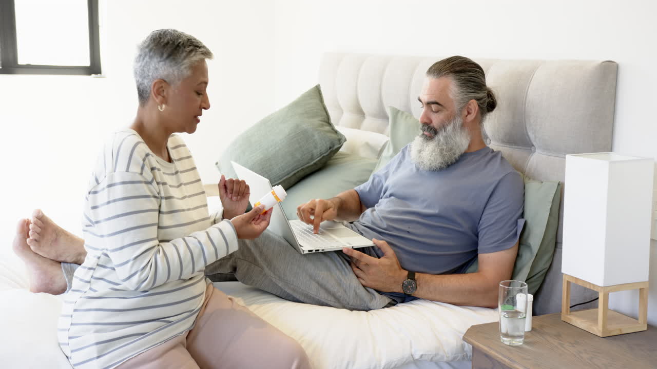 Senior couple discussing medication while using laptop in bedroom, relaxing together