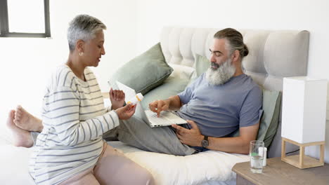 senior couple discussing medication while using laptop in bedroom, relaxing together
