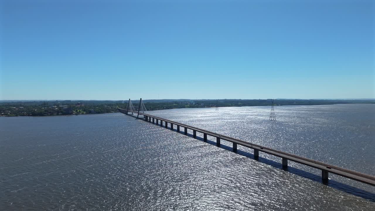 Panoramica aerea a ritroso che inquadra il Puente Internacional San Roque Gonzalez che attraversa un ampio fiume sotto un cielo azzurro e terso
