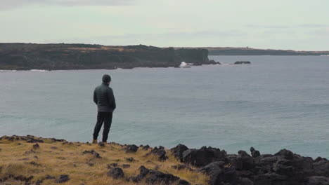 Man-stands-on-cliff-looking-out-at-the-waves-in-slow-motion