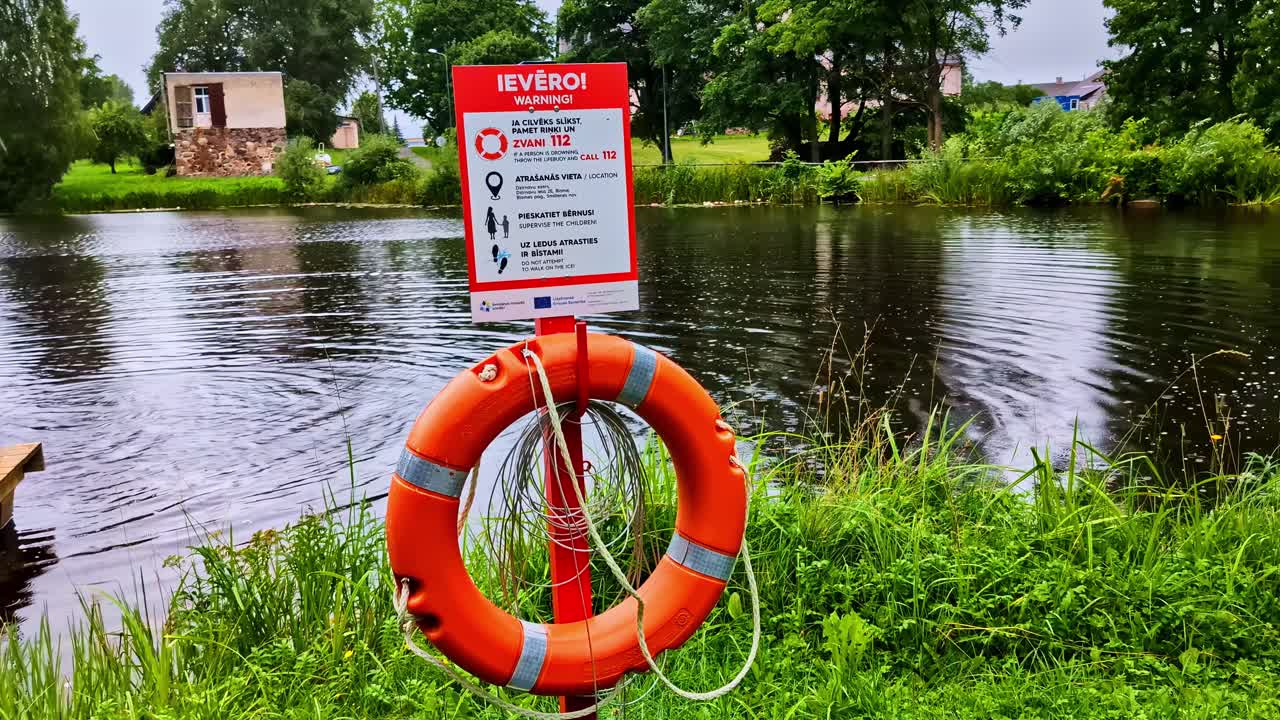 Bright Orange Lifebuoy With Warning Sign Near Calm Lake on Rainy Day
