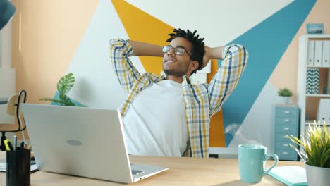 pleased afro-american man sitting at desk in office relaxing with hot drink on table