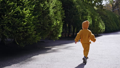 following the happy kid running by the road. baby boy in yellow suit and cap outdoors on sunny weather.