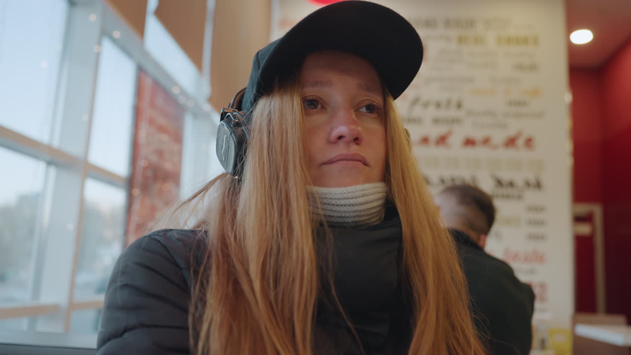 Donna bianca dai capelli rossi illuminata lateralmente che guarda fuori dalla finestra con le cuffie, pendolare silenziosa seduta in un caffè, espressione calma che oscilla tra stanchezza e riflessione, luce interna soffusa, sciarpa e berretto visibili.