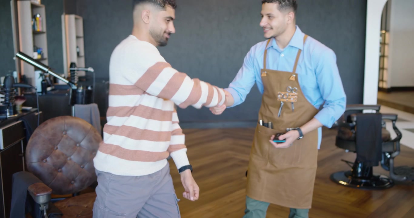 Cliente uomo e barbiere che si scambiano il telefono, il cliente che tocca lo schermo, controllando il taglio di capelli, uscendo dal negozio.