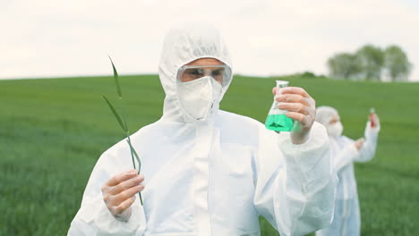 close-up view of caucasian researcher man in protective suit holding test tube and tree branch while doing pest control in the green field