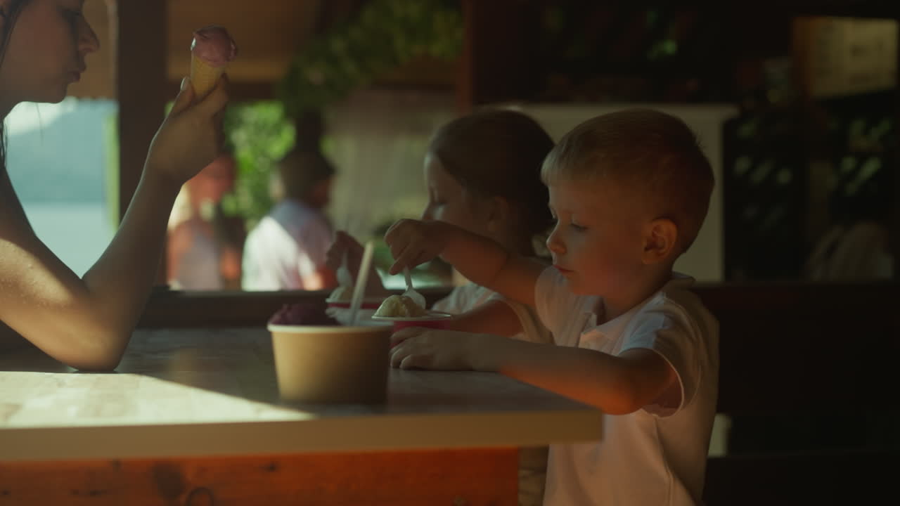 Toddler boy eats ice cream sitting with sister and mother in cafe slow motion. Happy family tastes cold desserts visiting restaurant. Summer lunch