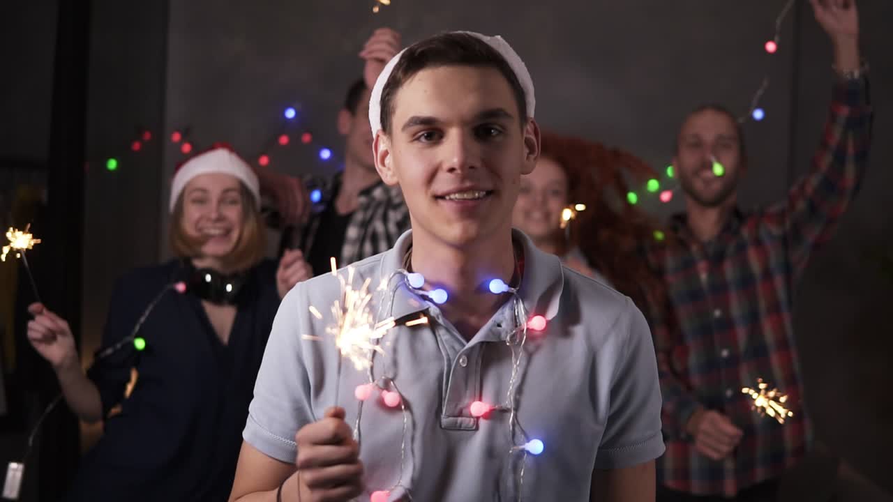 Portrait of caucasian young guy with colorful lights on neck and santa hat posing for camera - smiling, holding his bengal light while his friends dancing and celebrating on the blurred background in decorated room. Friends celebrating new year together