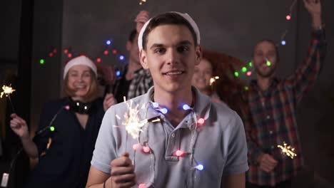 Portrait-of-caucasian-young-guy-with-colorful-lights-on-neck-and-santa-hat-posing-for-camera---smiling,-holding-his-bengal-light-while-his-friends-dancing-and-celebrating-on-the-blurred-background-in-decorated-room.-Friends-celebrating-new-year-together