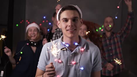 portrait of caucasian young guy with colorful lights on neck and santa hat posing for camera - smiling, holding his bengal light while his friends dancing and celebrating on the blurred background in decorated room. friends celebrating new year together