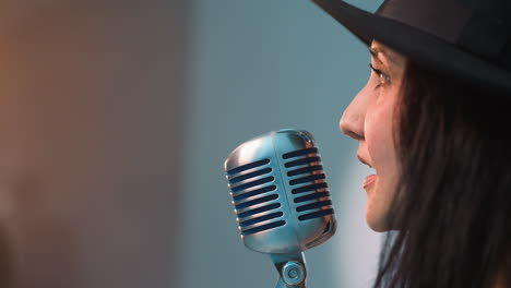 close side view of a singer wearing a black hat and holding a vintage microphone against a blue background. singer's facial profile and expression as she performs with focus and passion