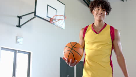 young biracial man with curly hair holds a basketball in a gym