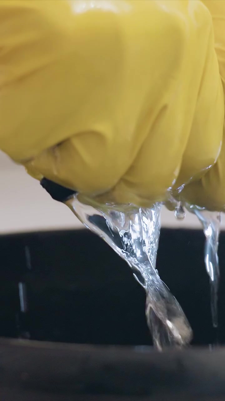 Shows yellow gloved hands repeatedly dipping into black bucket filled with clear liquid emphasizing material handling and manipulation without revealing full body or setting details