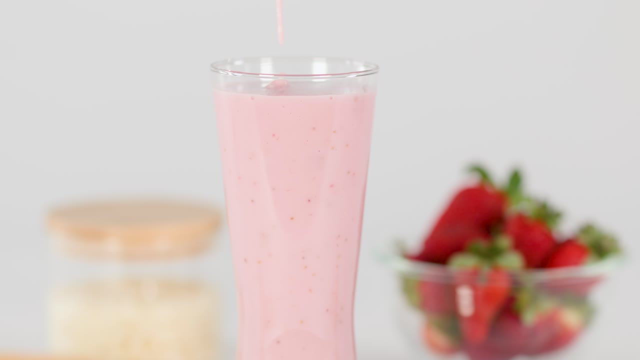Thick pink strawberry milkshake poured into glass, bright lighting, minimal background, smooth camera angle