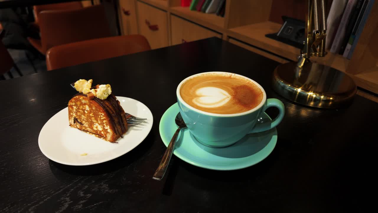 A cozy café setting with a slice of layered cake topped with cream on a white plate and a cup of cappuccino featuring latte art, placed on a dark wooden table.