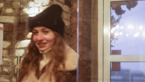 lady wearing black knit cap, brown shearling jacket, opening glass door and walking inside cafe on cold, overcast day, reflecting rainy weather outside