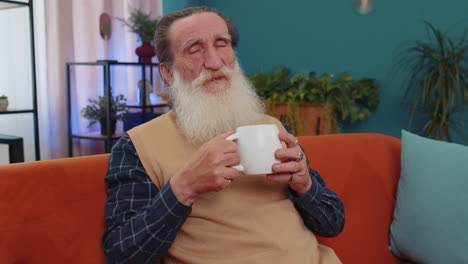 Attractive-smiling-senior-old-grandfather-man-drinking-a-cup-of-coffee-or-herbal-tea-at-home-couch