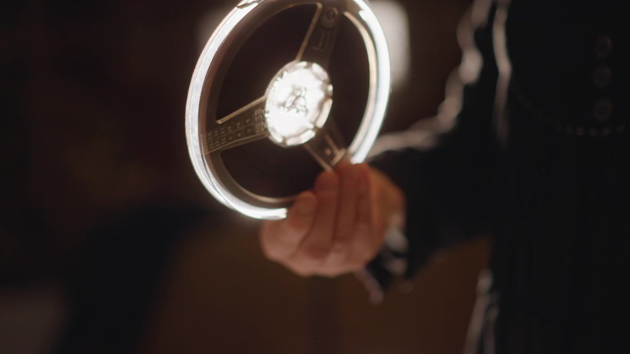 Persona che esamina minuziosamente una pellicola. Scatto dettagliato che mostra mani che tengono con cura una pellicola luminosa. Scena intima con mani che esaminano delicatamente una bobina di pellicola luminosa in una luce calda.
